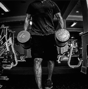 Black and white photo of Brandon performing a set of farmer's walks with a pair of 90 lb. dumbbells. 