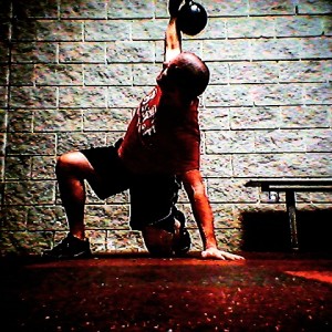 Brandon performing the Turkish Get Up exercise with the kettlebell in his right hand and his left knee and hand braced on the ground. 