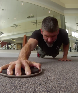 Brandon performing a glide disc push-up with his right hand on the disc sliding along the ground above his head. 