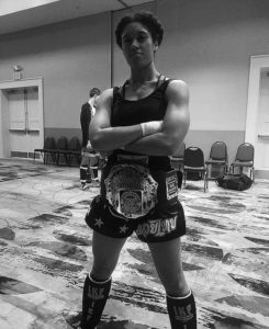 Black and white image of woman showing off a championship belt with her arms crossed posing for the camera. 