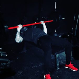 Dark image of a man performing a barbell bench press with a red barbell. 
