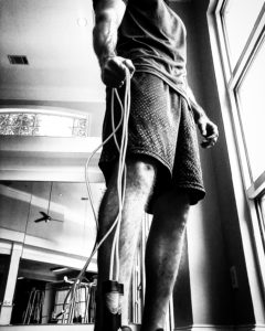 Black and white image of a man gripping the end of a jump rope with hand with it dangling by his side. 