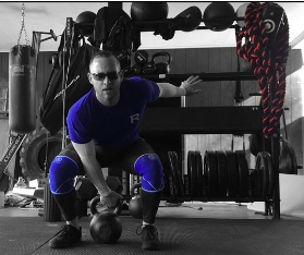 A man in blue with a gray background reaching down to grab a kettlebell for a clean and rack lift. 