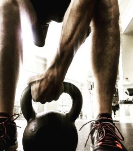 A kettlebell at ground level on the floor between the feet of a lifter with the lifter's muscular right arm grabbing the handle. 