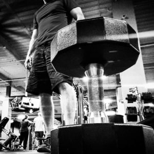 Black and white image of a dumbbell in the foreground of the image with a man standing in the background. 