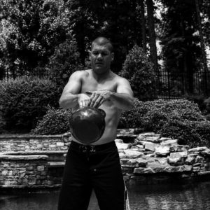 Black and white photo of Brandon performing a single arm kettlebell swings transitioning the kettlebell from one hand to the other. 