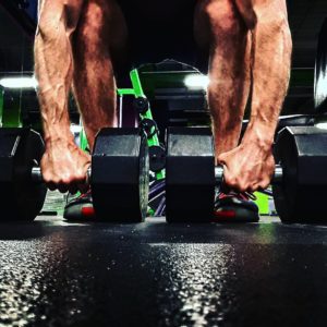 Man with muscular arms bending down to lift a pair of heavy dumbbells off the floor. 