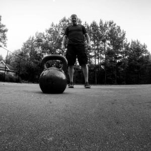 Black and white image with a kettlebell in the foreground and a man standing in the background. 