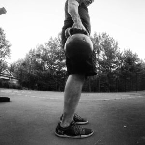 Black and white image of man standing with his right side to the camera holding a kettlebell in his right hand. 