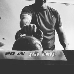 Black and white image of a man about to lift a kettlebell off of a box with his right arm. 